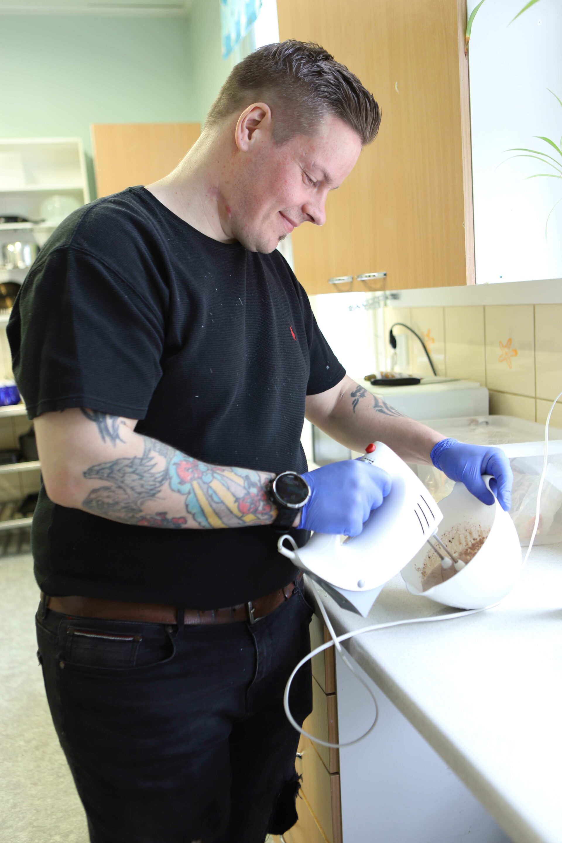 Hymyilevä lyhyttukkainen mies, jolla on farkut, teepaita ja tatuoitu käsivarsi, vatkaa sähkövatkaimella taikinaa keittiötason edessä.