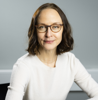 Polkkatukkainen, silmälasipäinen nainen, jolla on v-aukkoinen pusero.