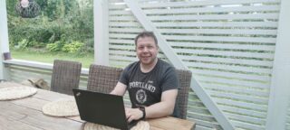 Hymyilevä, lyhyttukkainen mies istuu ulkona puutarha-aidan edessä ja kirjoittaa läppärillä.