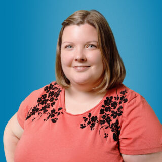 Vienosti hymyilevä polkkatukkainen nainen, jolla on jakaus keskellä päätä ja o-aukkoinen teepaita, jossa on olkapäillä kukkakuvioita.