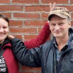 Tiiliseinän edessä seisoo vasemmalla hupparissa poninhäntäpäinen hymyilevä nuorehko nainen. Hän näyttää voitonmerkkiä vieressään oikealla puolella seisovan miehen pään päällä. Miehellä on lippalakki ja talvianorakki ja kauluspaita ja hän kättään naisen olkapäällä.
