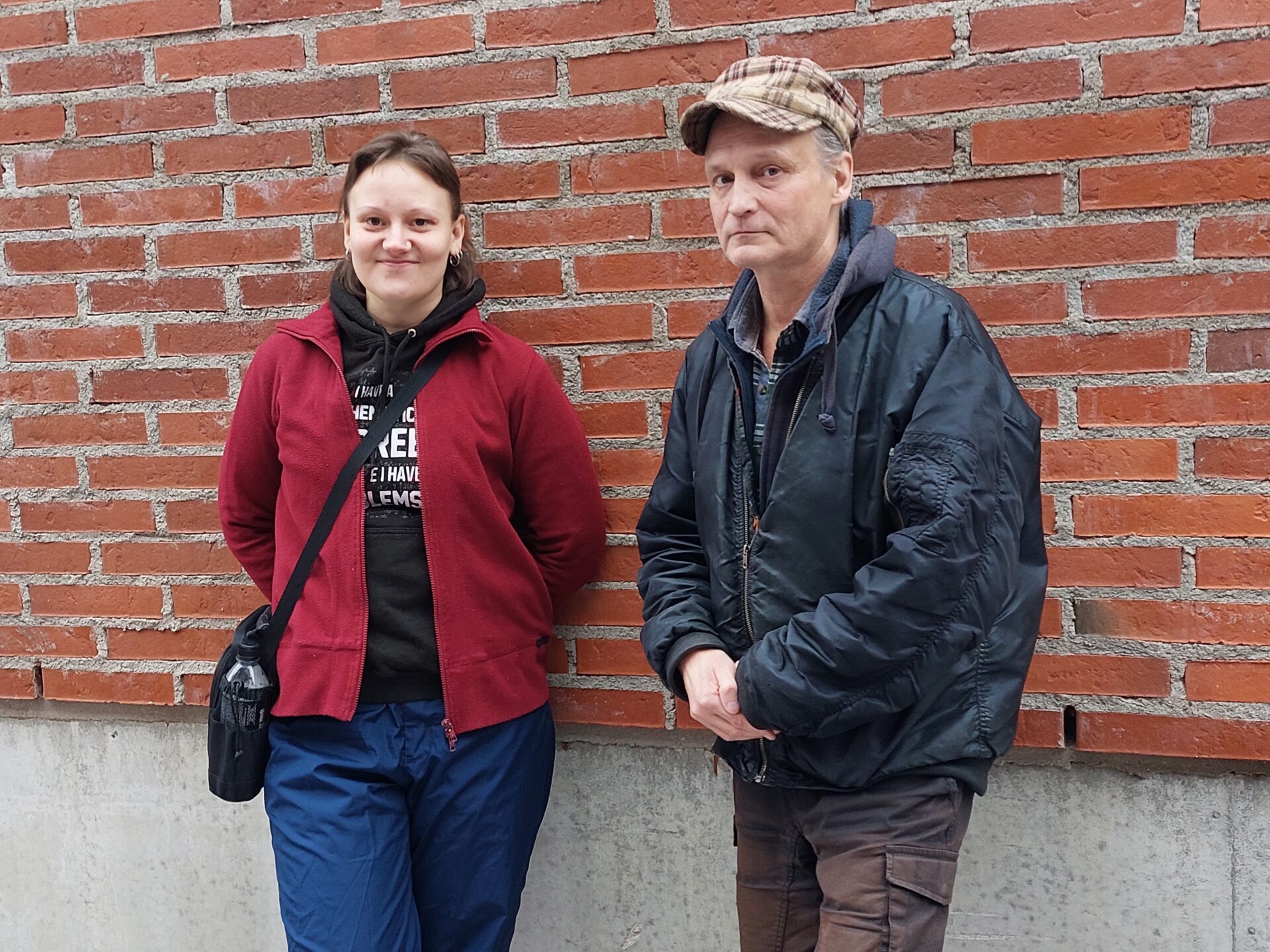 Tiiliseinän edessä seisoo vasemmalla hupparissa poninhäntäpäinen hymyilevä nuorehko nainen. Oikealla on vakavahko lippalakkipäinen mies, jolla talvianorakki ja kauluspaita.