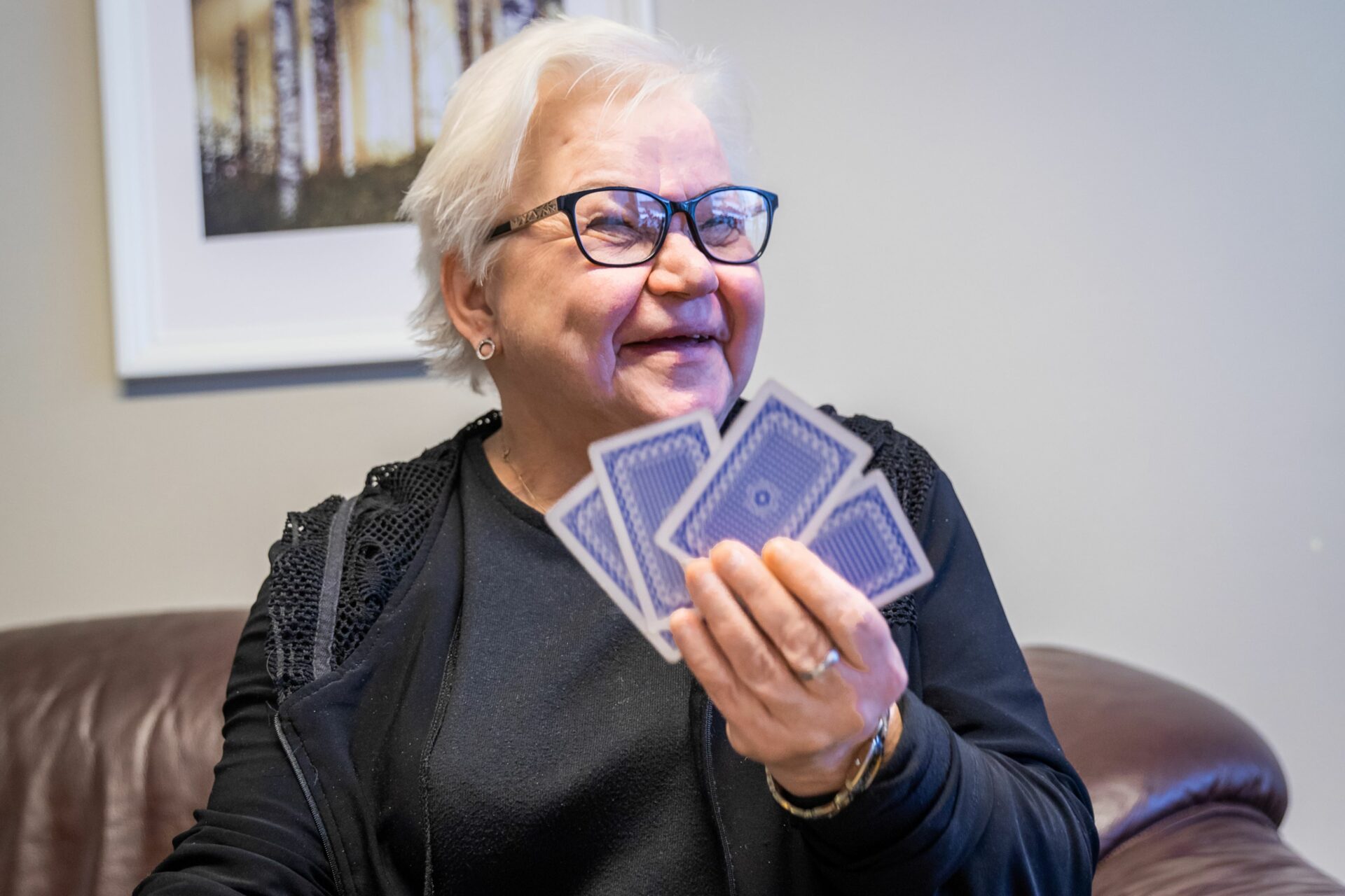 Lyhyttukkainen,silmälasipäinen, iloisesti hymyilevä nainen pitää kädessää viuhkana pelikortteja.