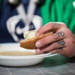 Tatuoidussa kädessä on leipäpala, jolla on kurkkua. Käden takana on keittolautanen. Taustalla on ihmishahmo.