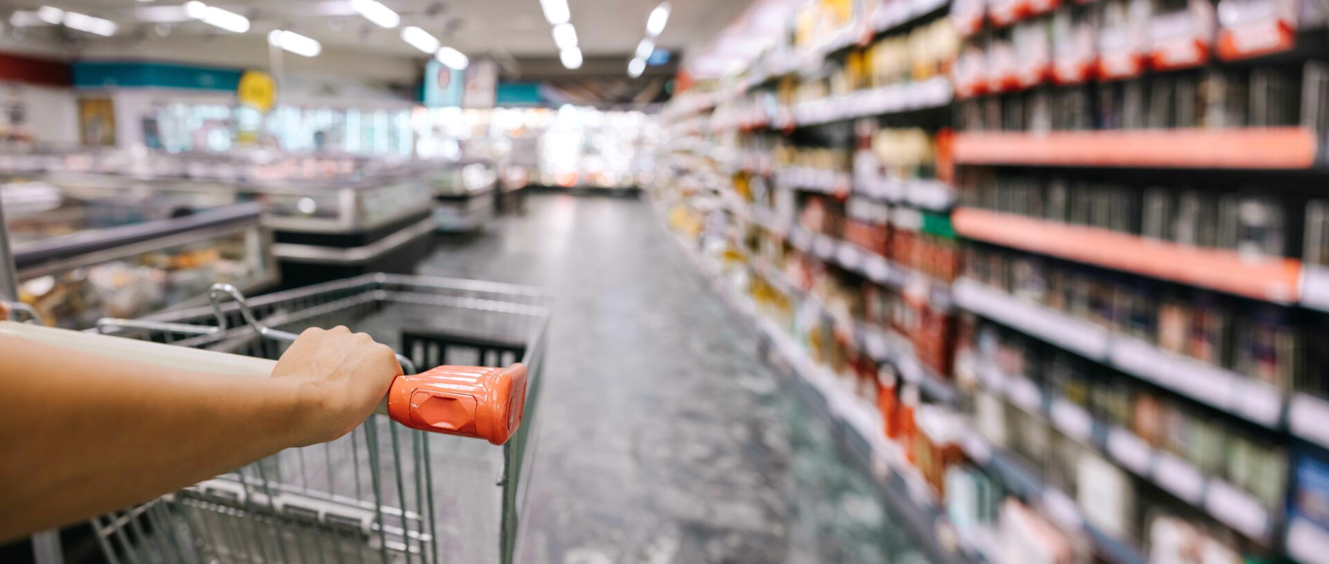 Etualalla ostoskärryistä kiinni pitävä käsi. Taka-alalla marketin käytävä.
