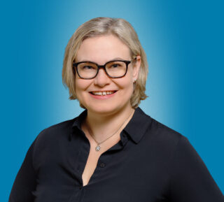 Lyhyttukkainen, silmälasipäinen hymyilevä nainen, jolla on rako etuhampaiden välissä. Hänellä on kauluksellinen paitapusero ja kaulassa ohut koru,