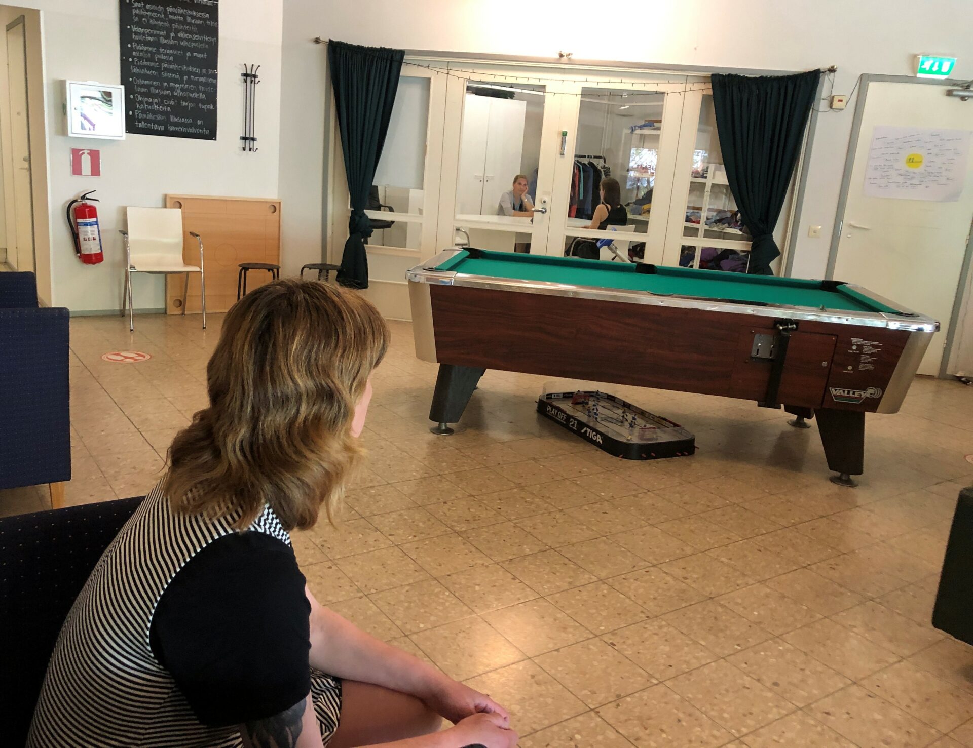 Takaapäin pitkätukkainen henkilö, joka istuu sohvalla. Taustalla on biljardipöytä, jonka alla on fortunapeli, ja iso neljäosainen ikkuna, jossa on maahan asti ulottuvat verhot.