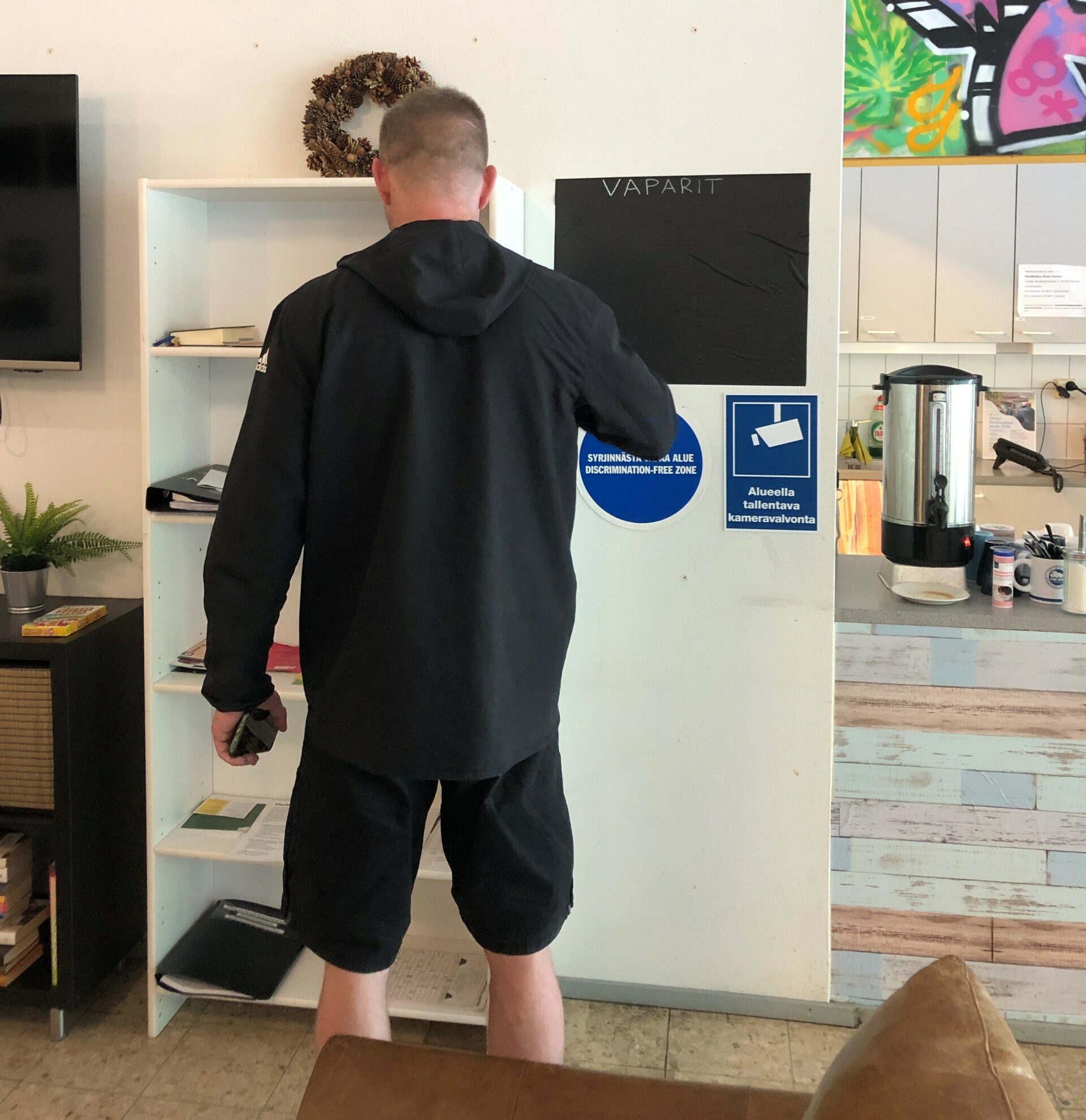 Takaapäin hupparipukuinen mies, joka seisoo matalan hyllyn edessä. Taustalla on keittiötila.