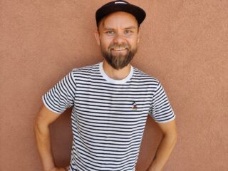 Hymyilevä, parrakas mies, jolla on lippalakki ja raidallinen teepaita.