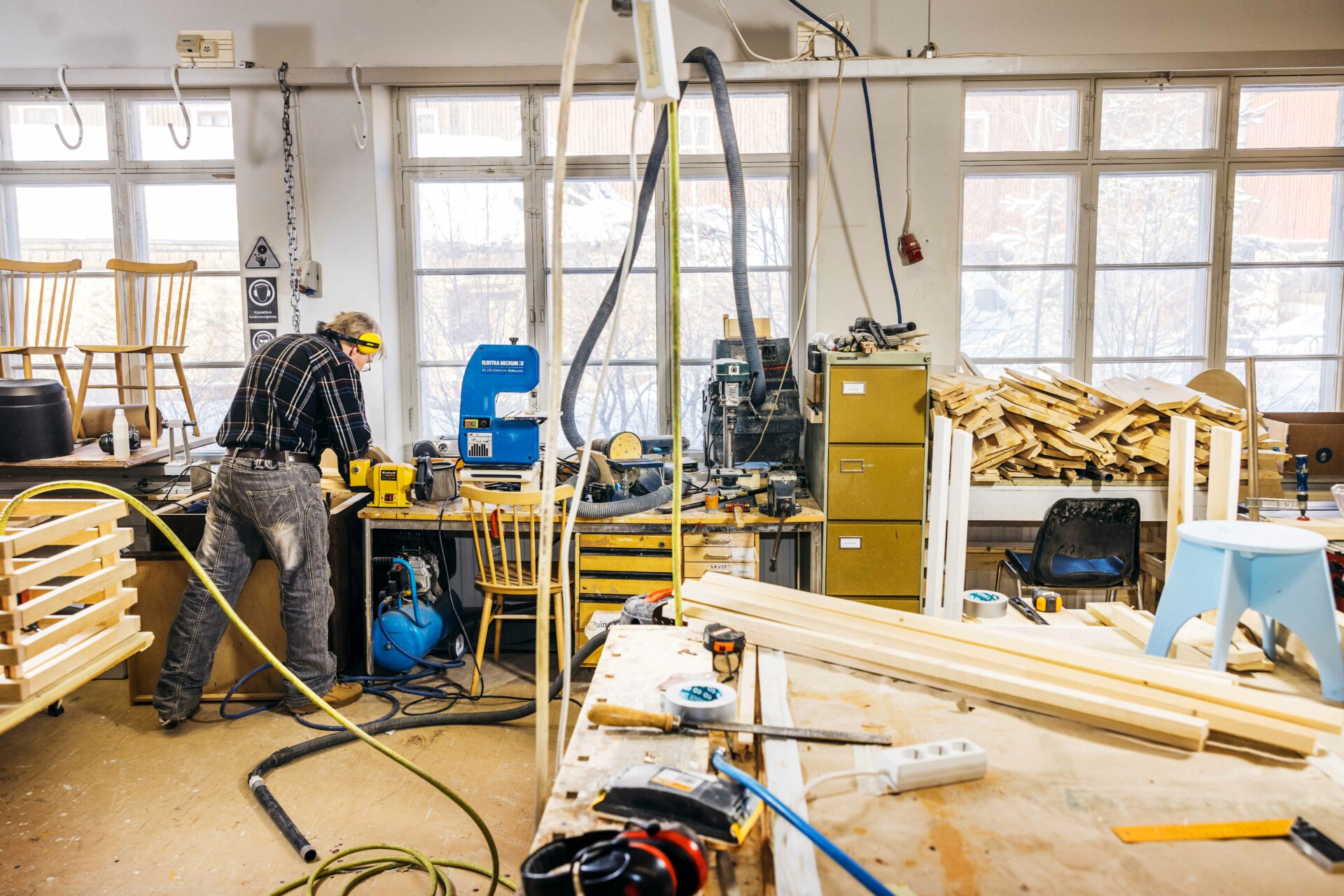 Puuverstaalla työskentelee noin 60-vuotias mies, selin, kasvoja ei näy. Työkoneiden äärellä.