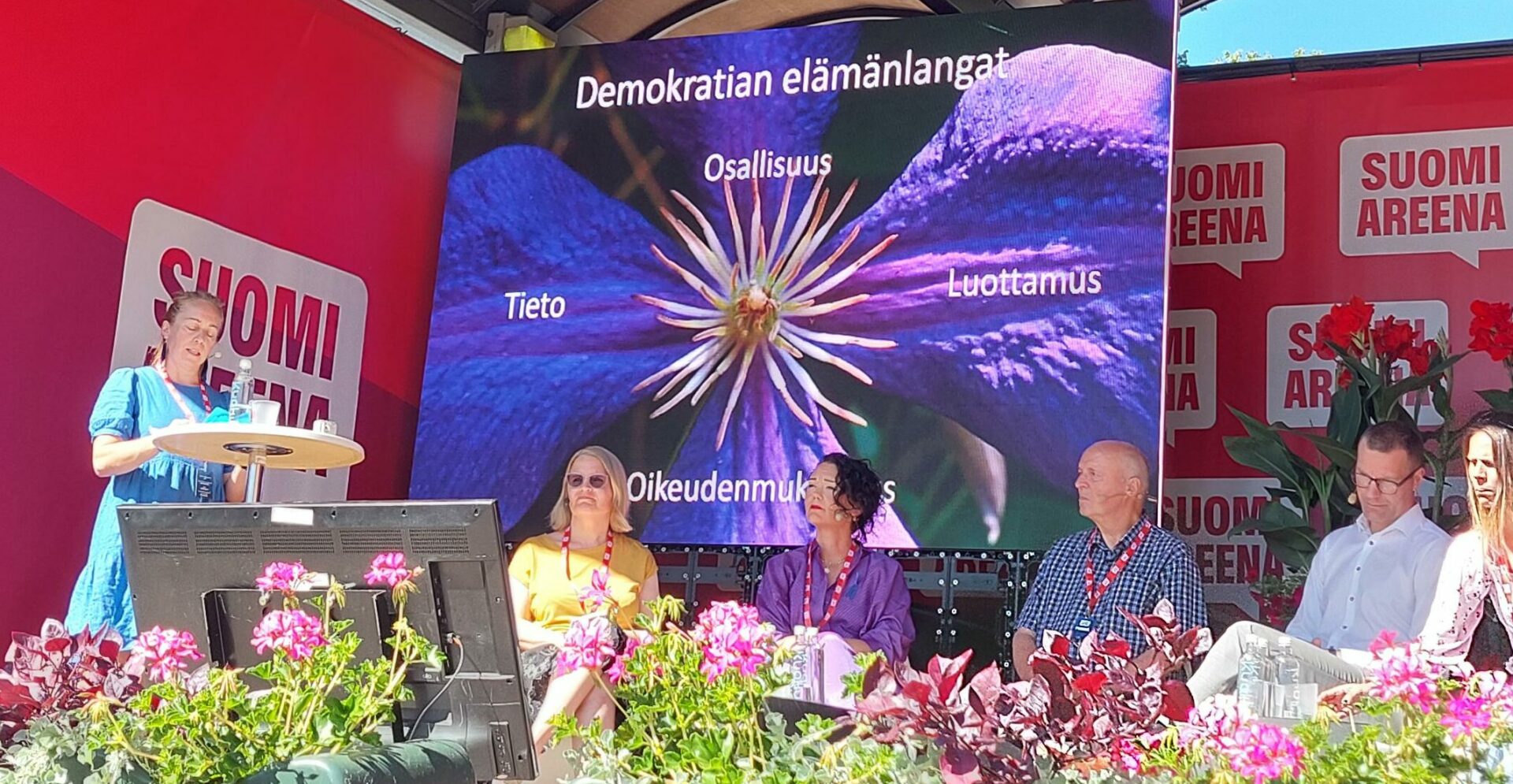 Porissa SuomiAreenan lavalle keskustelutilaisuudessa juontaja puhuu ja kolme ihmistä istuvat lavalla. Taustalla kuva demokratian säikeistä.