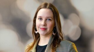 Pitkätukkainen, kapeakasvoinen, vienosti hymyilevä nainen, jolla on bleiseri ja o-aukkoinen paita. Tausta on sumennettu.