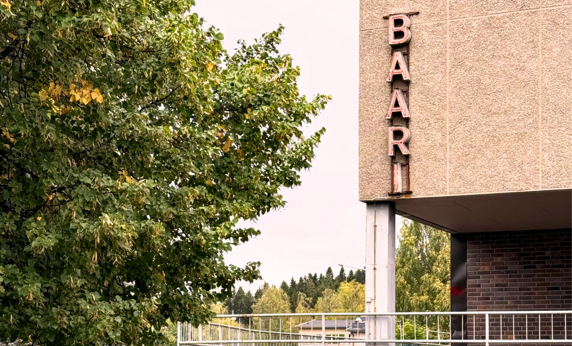 Vaalean ruskean kerrostalon reunassa Baari-kirjaimet, vaemmalla lehtipuu.