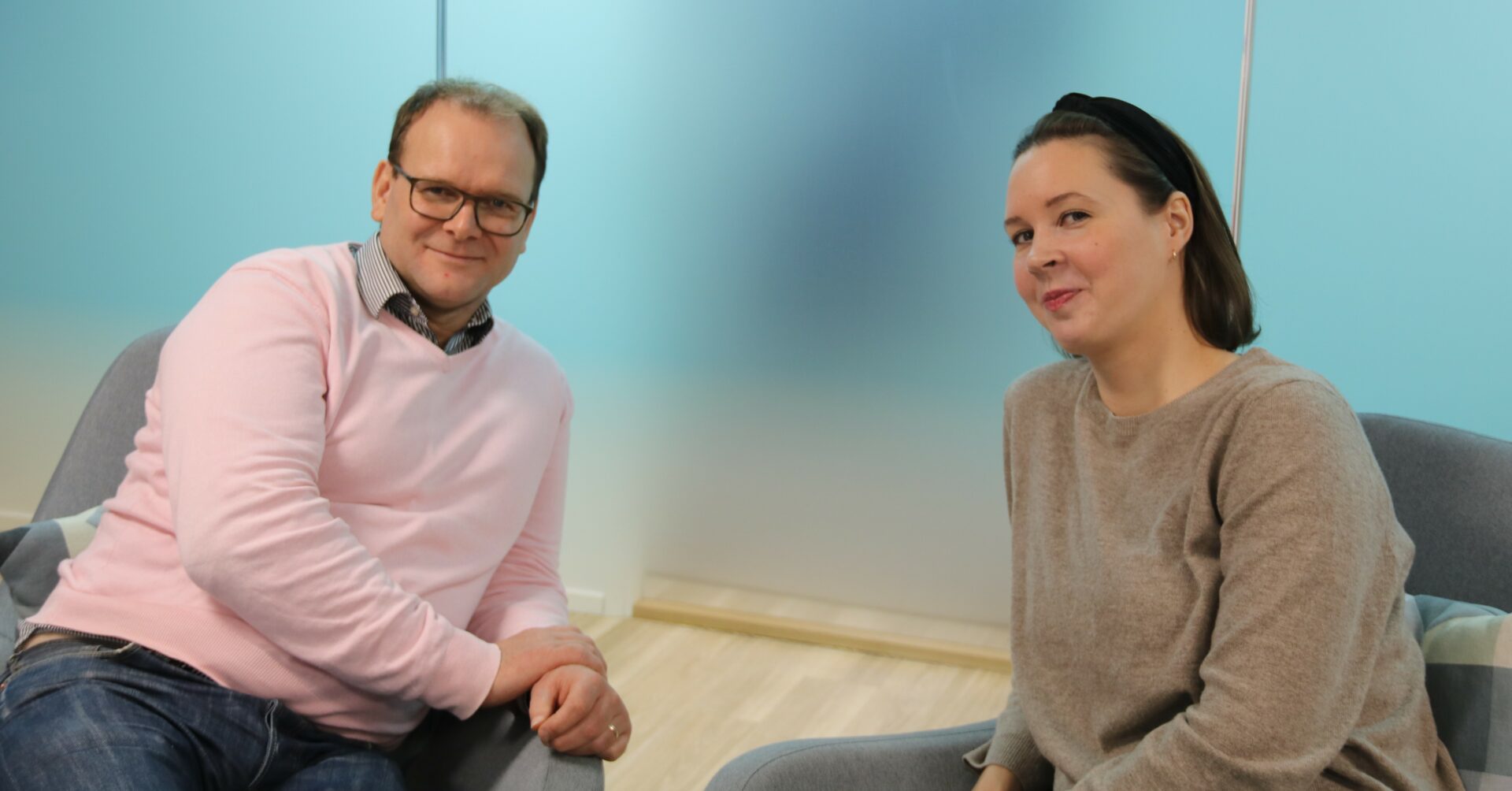 Vasemmalla hymyilevä, silmälasipäinen mies, jolla on neulepusero ja kauluspaita. Oikealla hymyilevä, polkkatukkainen nainen, jolla on hiuspanata ja neulepusero. Molemmat istuvat. Taustalla on läpinäkymätön lasiseinä.