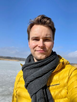 Noin 30-vuotias ruskeatukkainen mies katsoo kameraan. Päällä kaulaliina ja keltainen toppatakki. Taustalla taivas ja jäinen järvi.