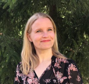Pitkätukkainen hymyilevä nainen, jolla on vee-aukkoinen mekko. Taustalla on lehvikköä.