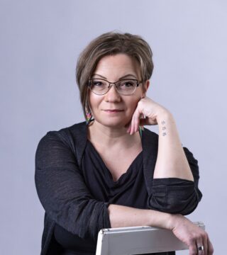 Ruskeatukkainen, keski-ikäinen näinen, jolla silmälasit katsoo kameraan. Vasen käsi poskella.