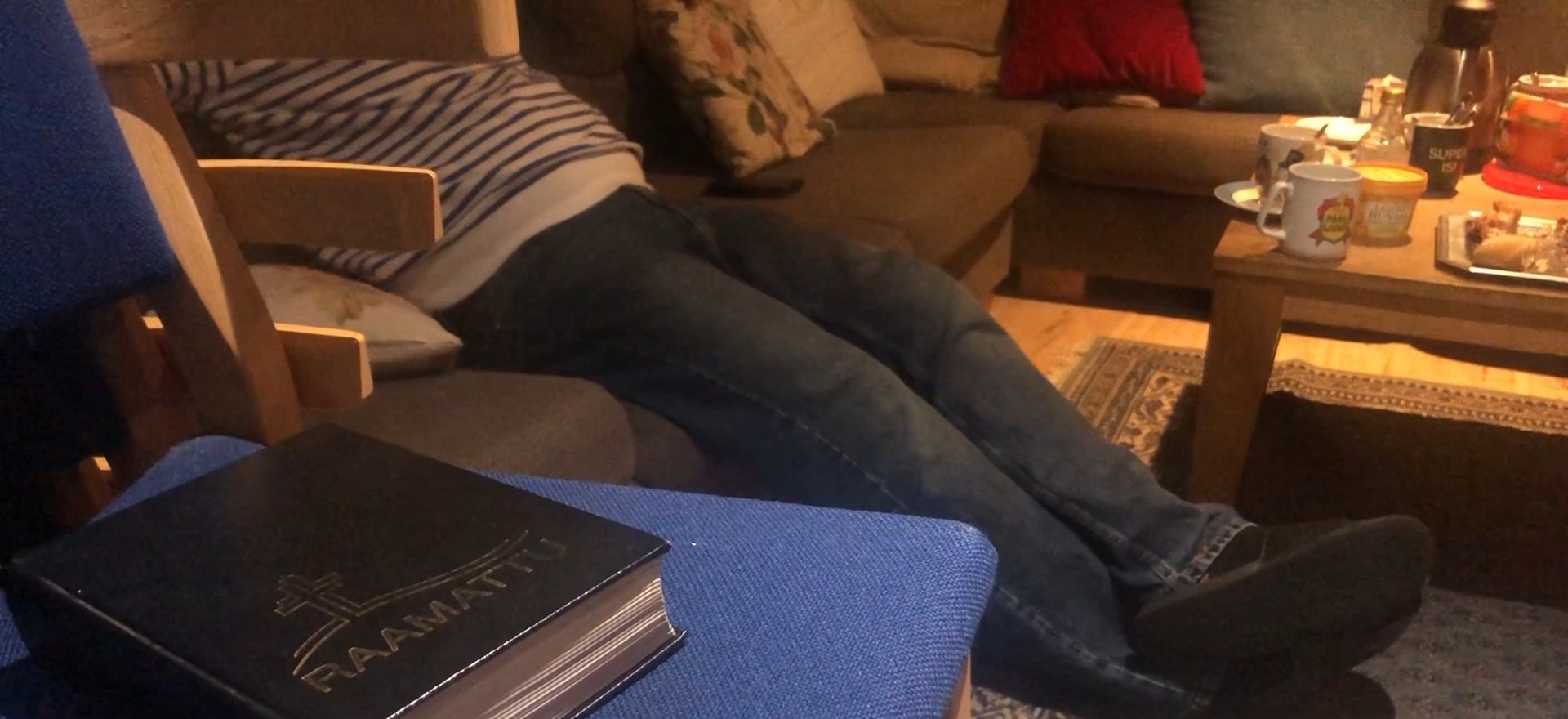 Lepotuoliryhmä ja sohvapöytä. Vasemmalla tuolissa istuu mies jalat pitkästi eteen ojennettuina. Etualalla tyolin päällä on Raamattu. Sohvapöydällä on kahvikuppeja.