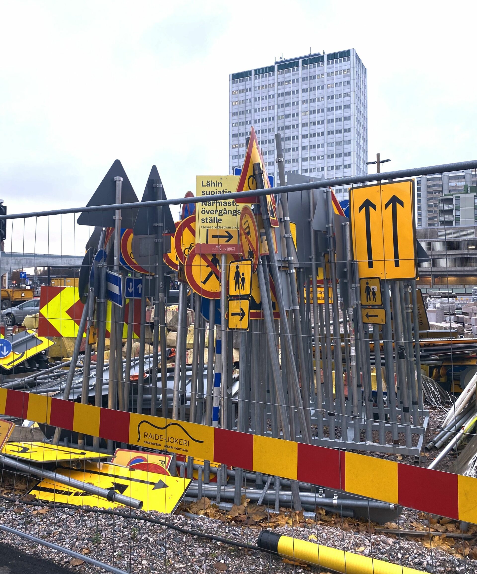 Sekalainen kasa liikennemerkkejä, jotka ovat sekä pystyssä että lappeellaan maassa. Taustalla on korkea rakennus.