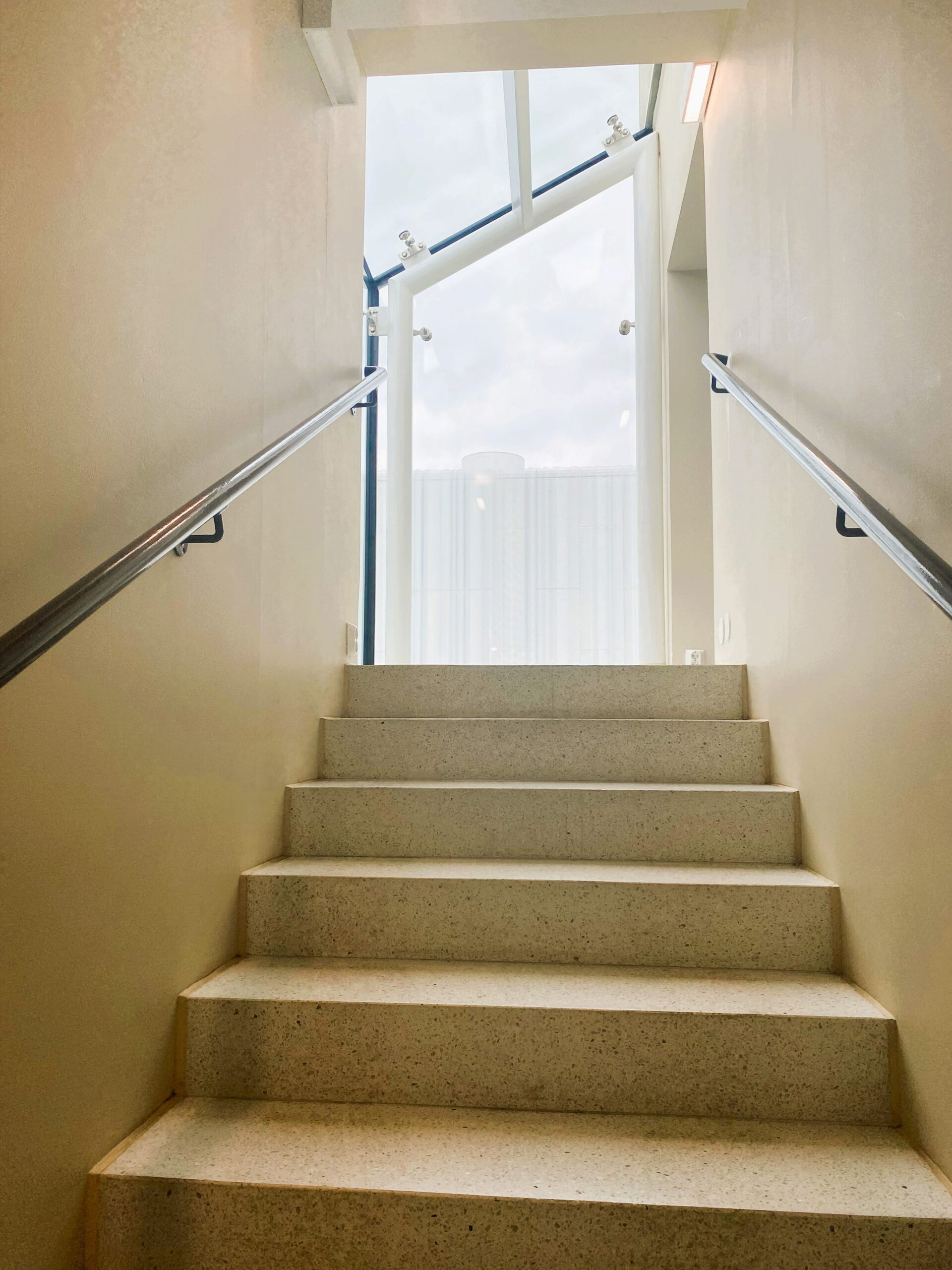 Ylöspäin suuntaavat portaat, joissa on kaiteet molemmin puolin. Portaiden yläpäässä näkyy valoisa ikkuna.