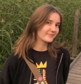 Pitkätukkainen, vienosti hymyilevä nuori nainen. Taustalla vihreää kasvistoa.