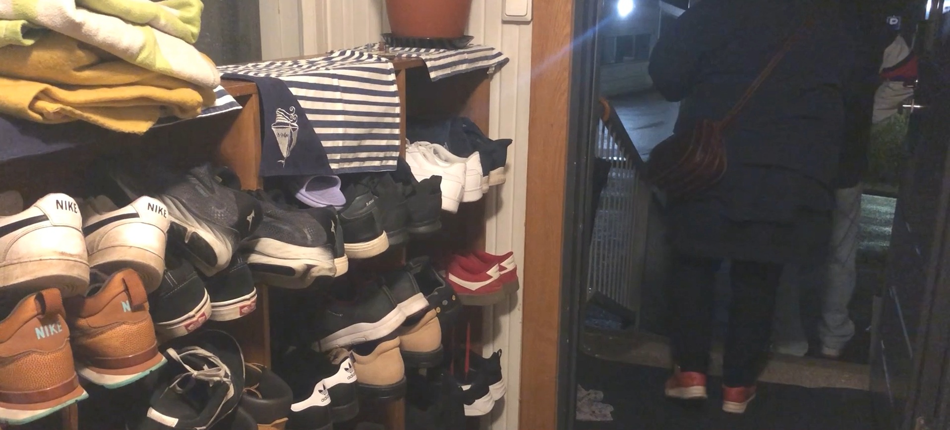 Eteistila, jossa vasemmalla on paljon kenkiä usealla hyllytasolla. Oikealla on kaksi ihmistä poistumassa avoimesta ovesta.