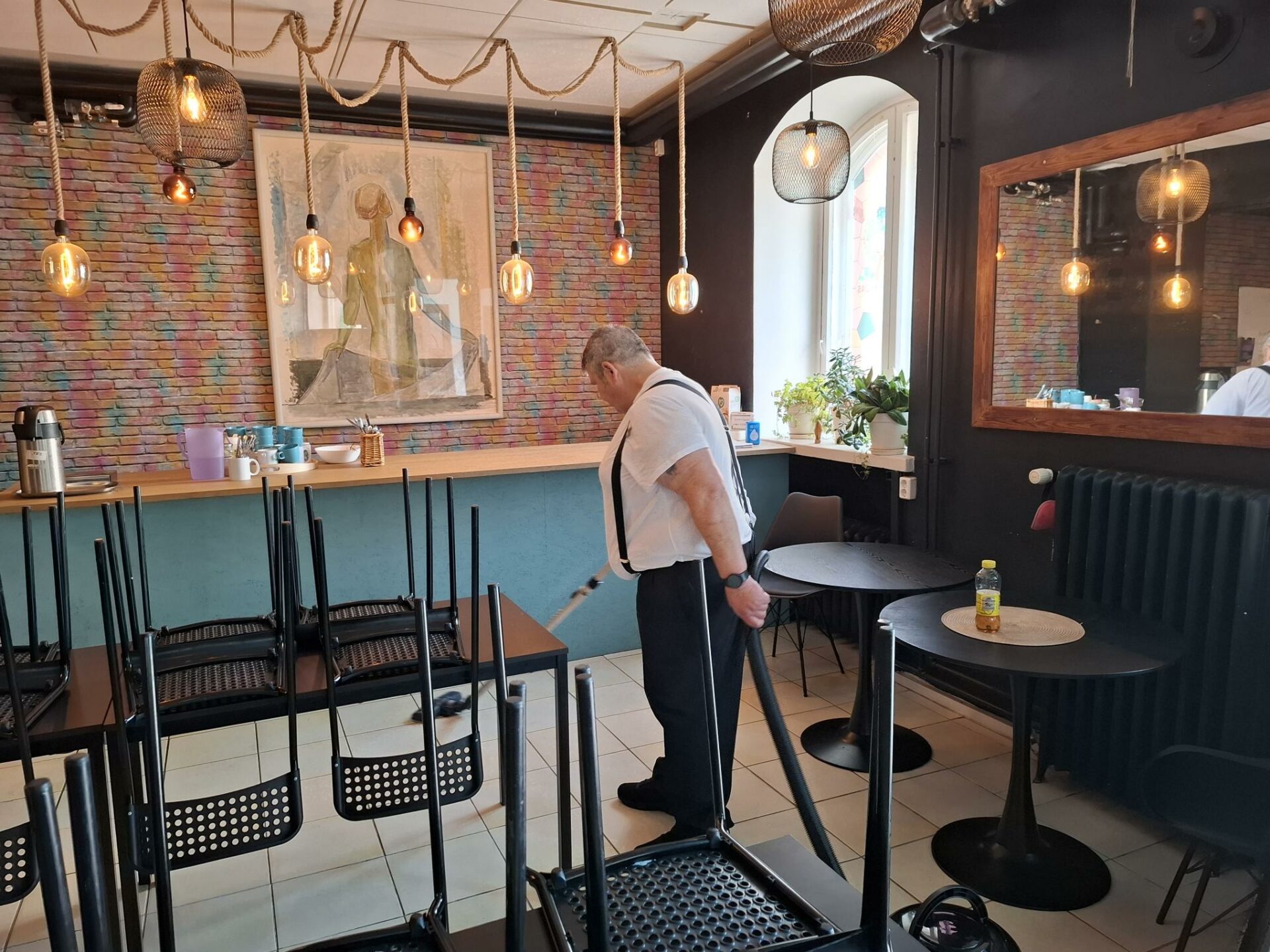 Paitahihasillaan oleva mies imuroi oleskelutilaa, jossa on vasemmalla pystypöytiä ja oikealla iso pöytä, jonka päällä on tuoleja. Taustalla on iso taulu ja ikkuna. Katosta riippuu pieniä valaisimia.