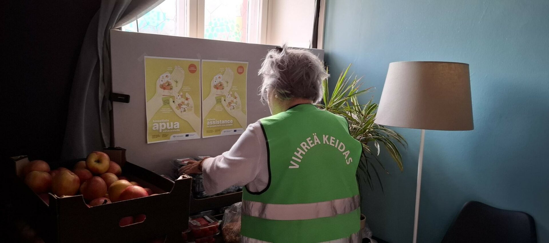 Lyhyttukkainen nainen selin. Taustalla ylhäällä on ikkuna, vasemmalla pöydällä omenoita ja oikealla jalkalamppu.