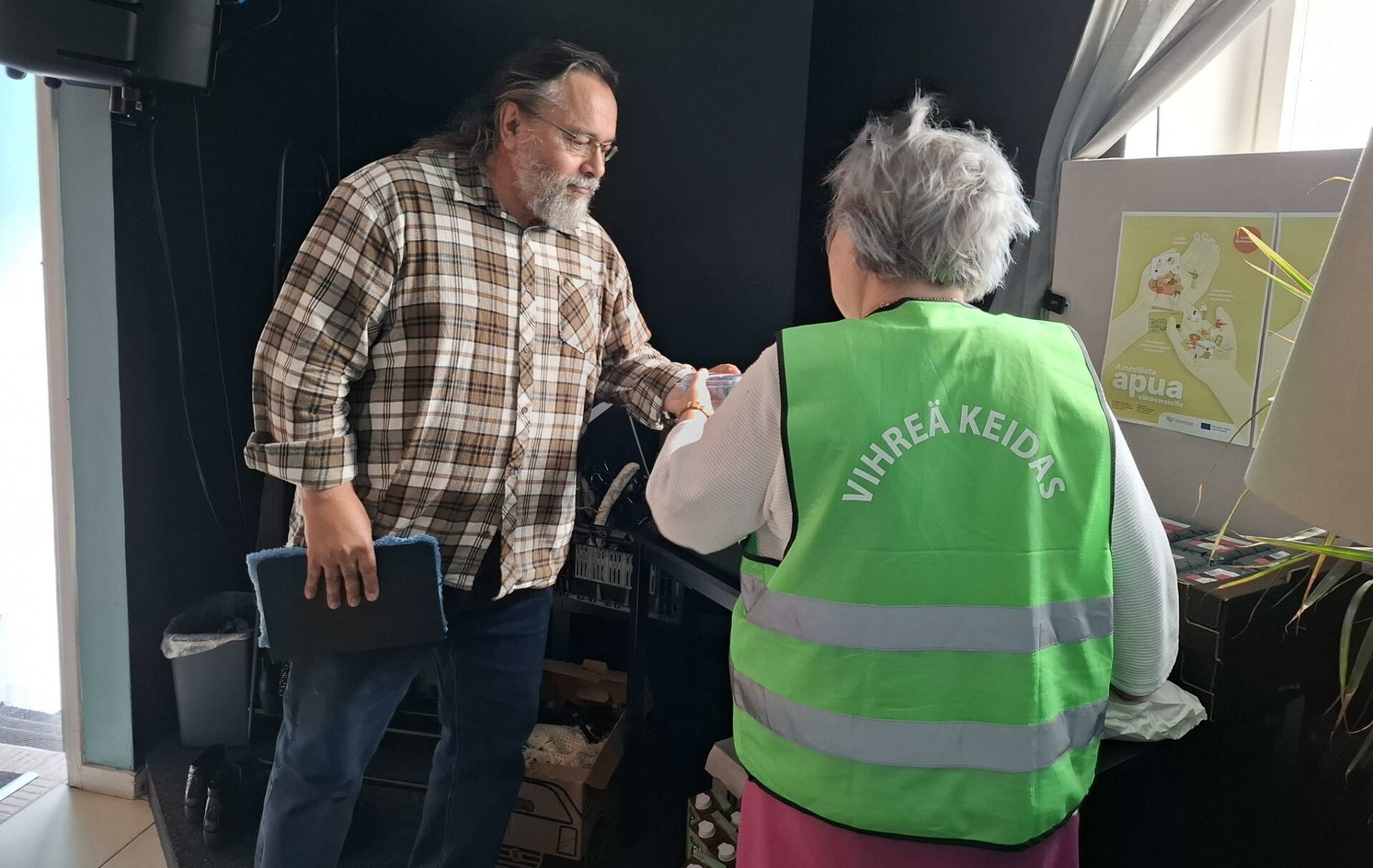 Parrakas mies ja selin lyhyttukkainen nainen keskustelevat. Naisella on Vihreän keitaan huomioliivi.