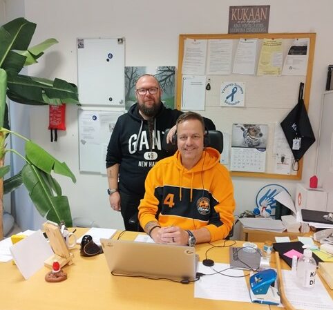 Oranssipaitainen, hymyilevä mies istuu kirjoituspöydän ja tietokoneen ääressä. Hänen takanaan seisoo tummaan huppariin pukeutunut silmälasipäinen, parrakas mies. Kirjoituspöydällä on paljon papereita. Taustalla on ilmoitustaulu.