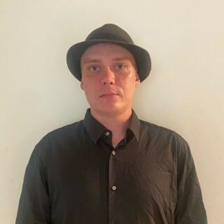 Nuorehko mies, jolla on lierihattu, kauluspaita ja vakava ilme.