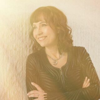 Kiharatukkainen nainen, jolla on otsahiukset, katsoo hymyillen vasemmalle. Hänellä on rennosti kädet puuskassa, nahkatakki ja kaulakoru.