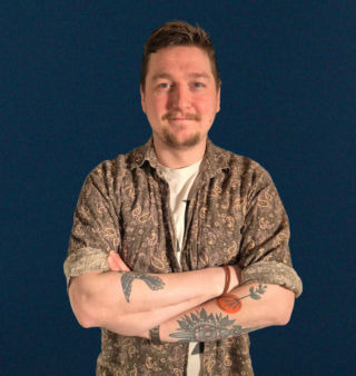 Miehellä on lyhyt pystytukka, pieni parta, teepaita ja sen päällä kuviollinen kauluspaita. Hänellä on käsivarsissa tatuointeja ja kädet ovat puuskassa rinnan päällä.