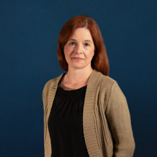 Pitkätukkaisella naisella on tumma teepaita ja neulostakki. Hänellä on kädet taskuissa.