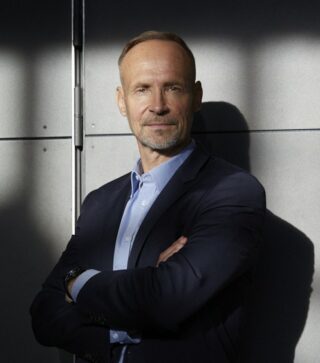 Sänkitukkainen, vakavahko mies kädet puuskassa. Miehellä on kauluspaita ja pikkutukka.