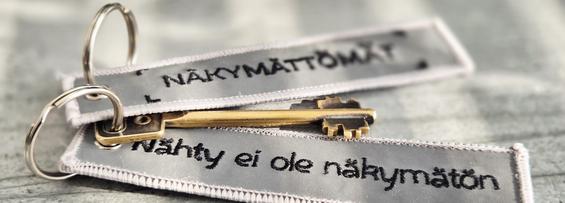 Avainrengas, jossa on avain ja kaksi kangasheijastinta, joissa toisessa on teksti Näkymättämät ja toisessa Nähty ei ole näkymätön.