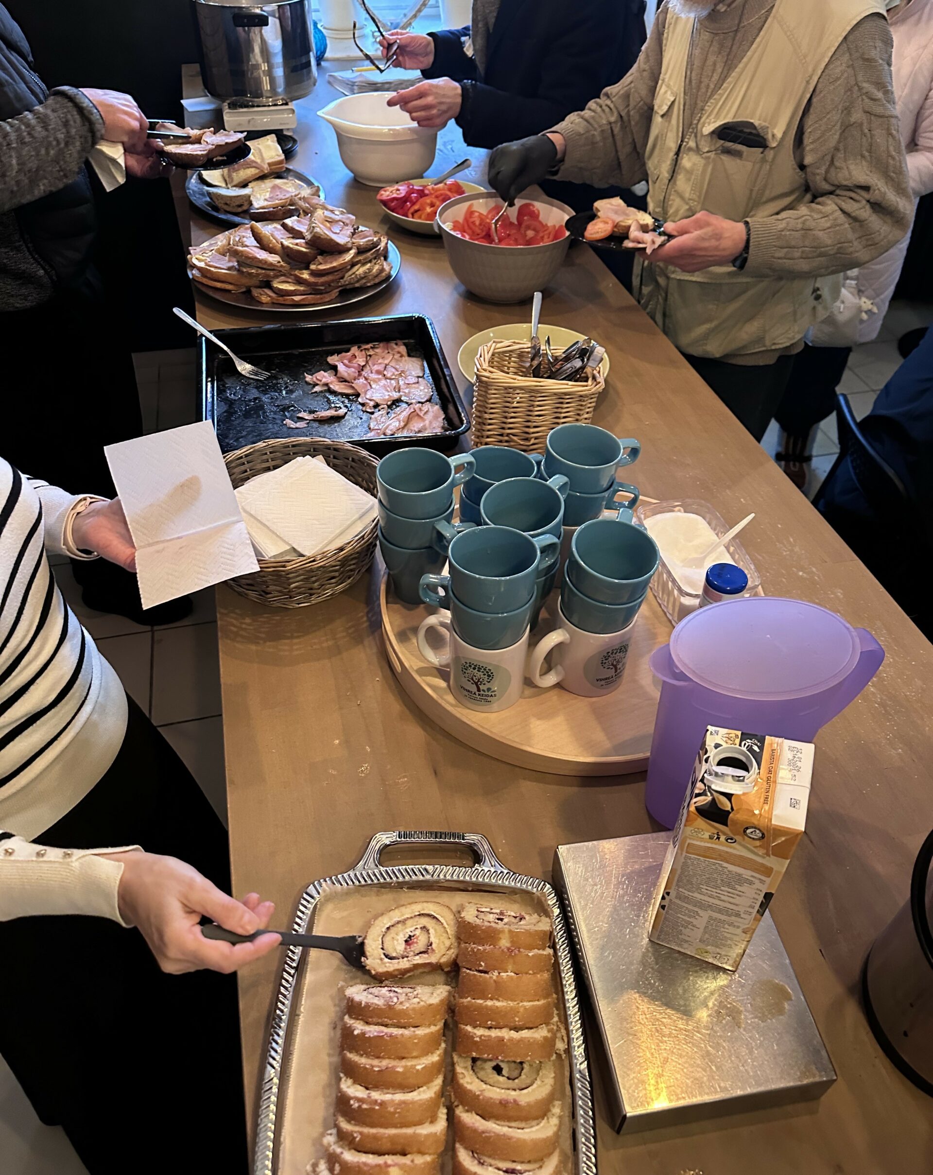 Pöydällä on kahvikuppeja ja kääretorttua. Ihmisten käsiä ojentuu ottamaan kuppeja ja tarjottavia.