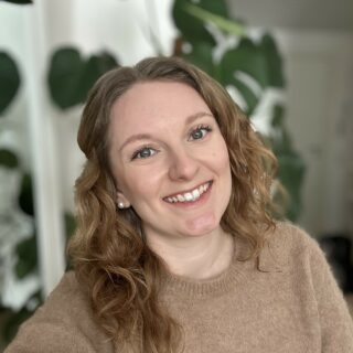 Leveästi hymyilevä nainen, jolla on pitkät kiharat hiukset ja yllään oo-aukkoinen neule. Taustalla kuviolliset verhot.