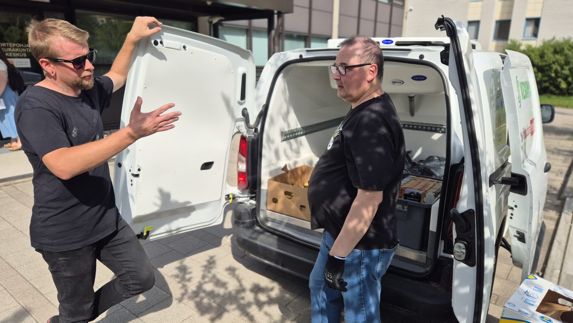 Kaksi miestä keskustelee pakettiauton takana. Pakettiauton takaovi on auki.