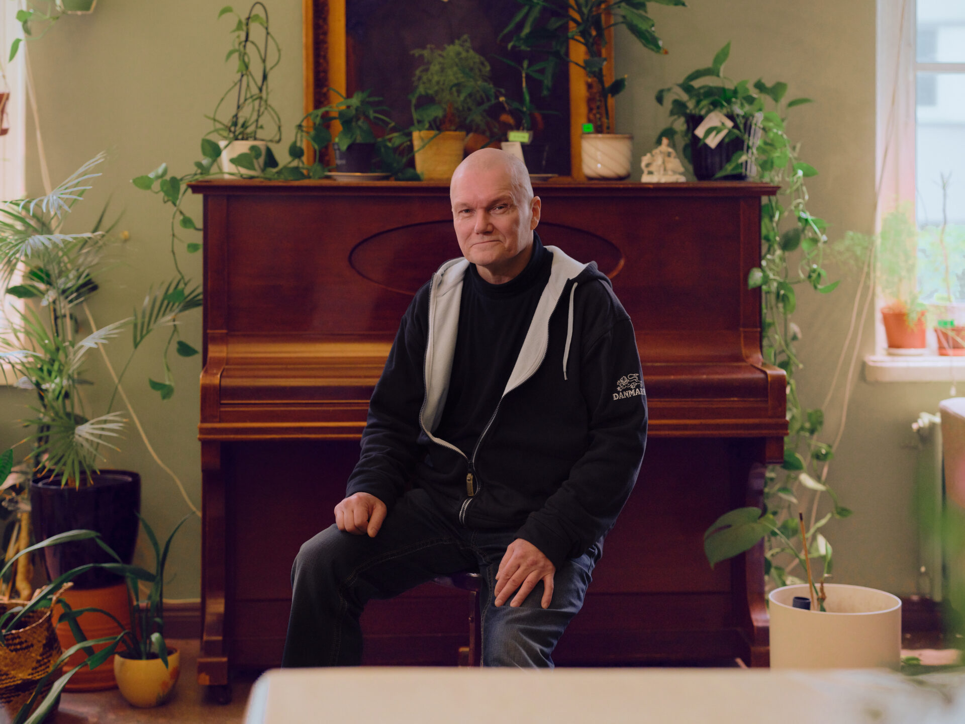 Hymyilevä, kalju mies istuu pianojakkaralla pianon edessä kädet polvien päällä.