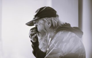 Iäkäs lippalakkipäinen mies, jolla on käsi kasvojen edessä.