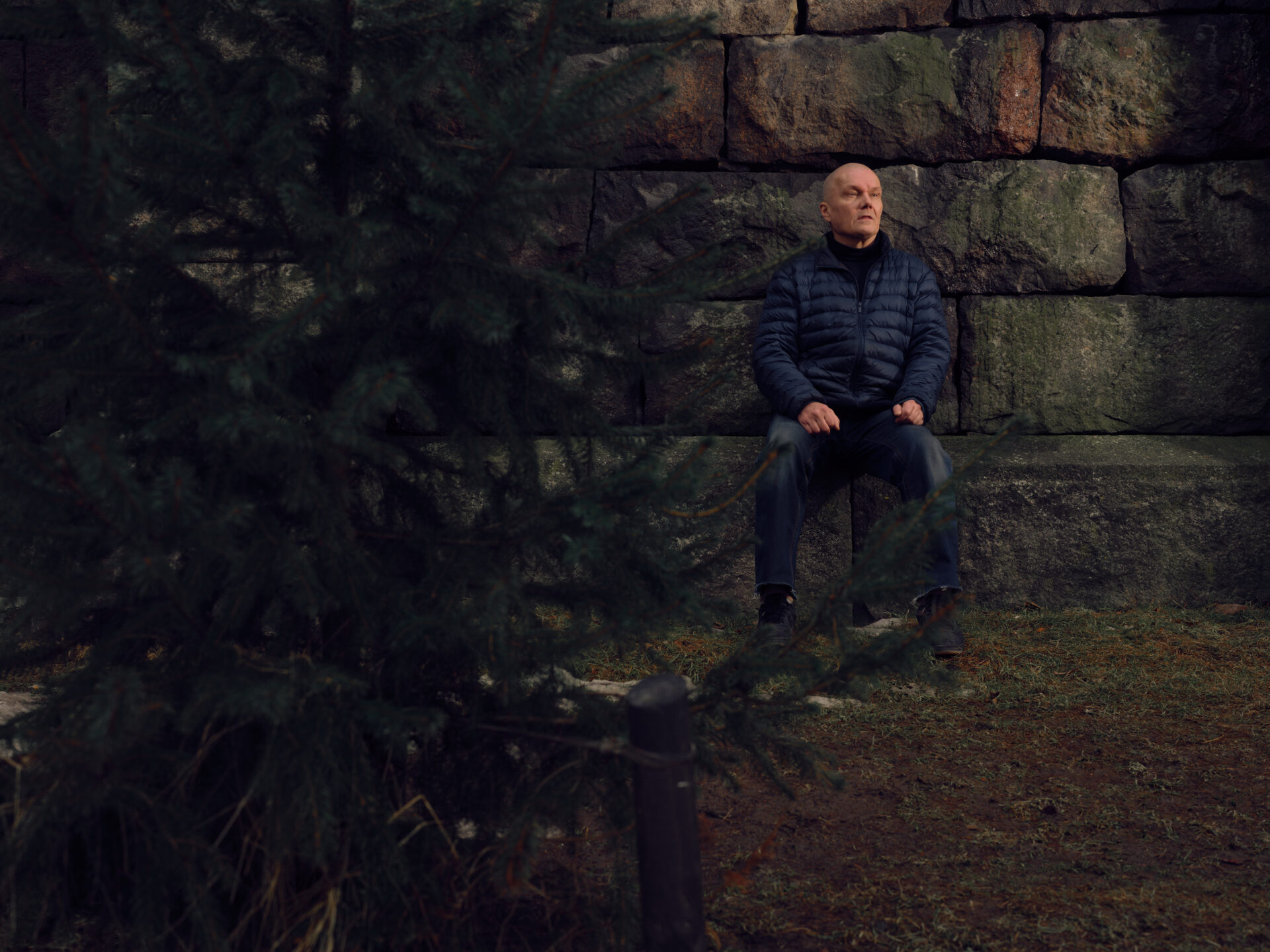 Vakava, kalju mies istuu kiveyksen reunalla miltei kuusen takana.