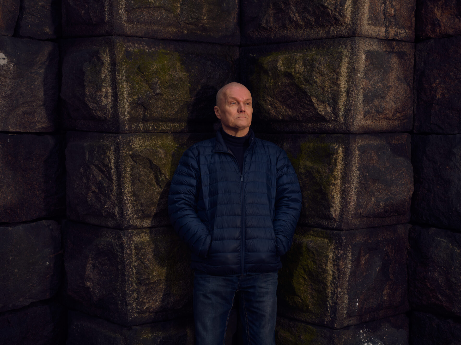 Vakava, kalju mies seisoo kädet takin taskuissa kivimuurin edessä.