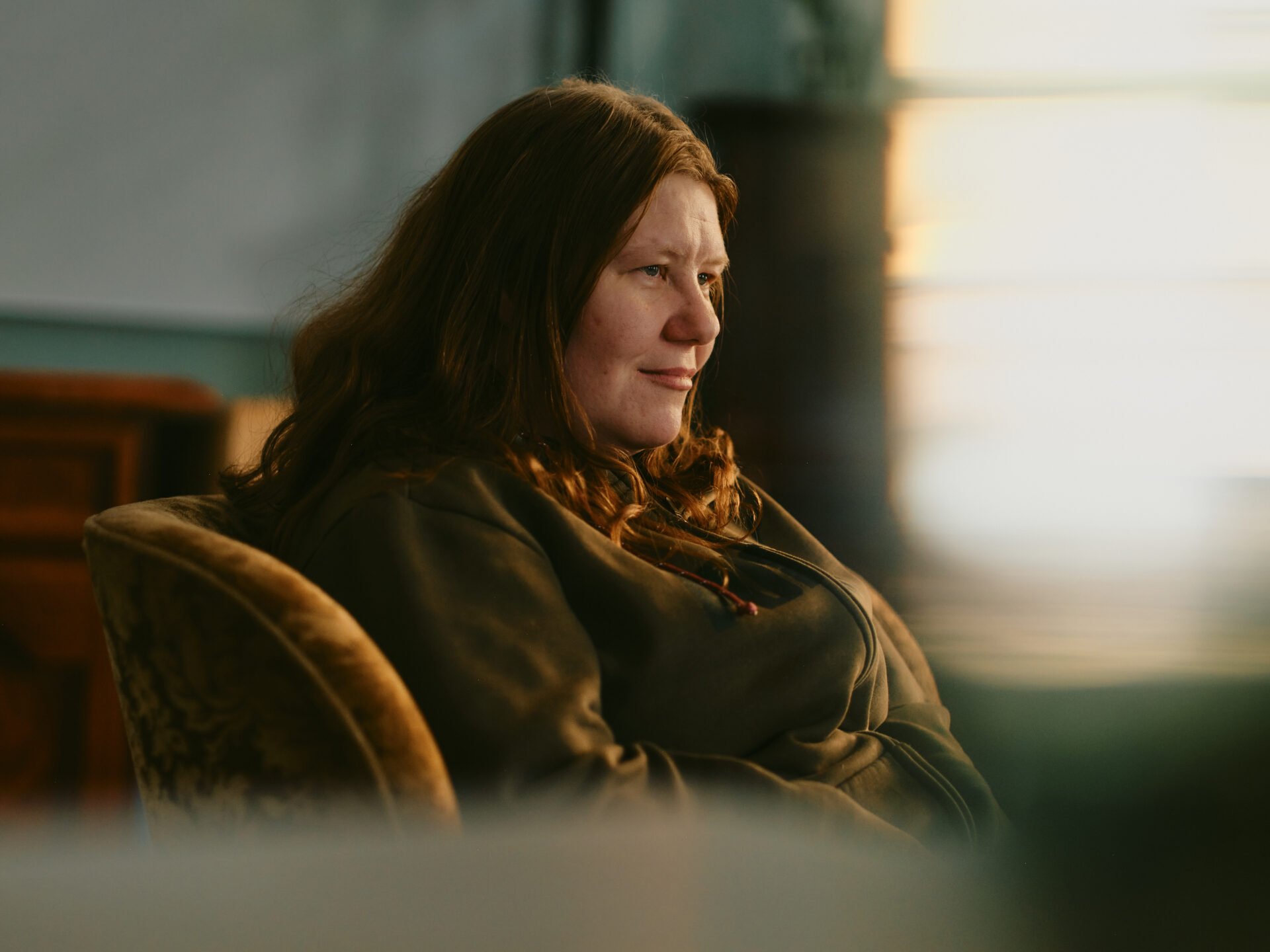 Pitkätukkainen nainen istuu nojatuolissa ja katsoo sivuun.