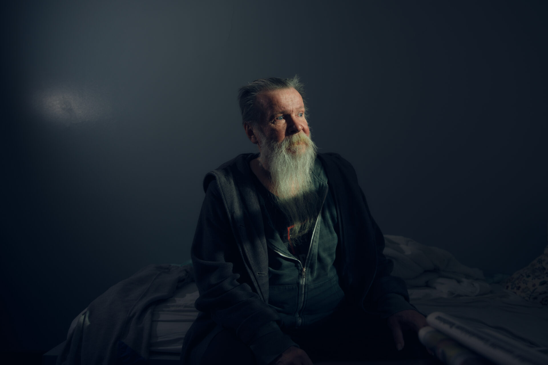 Iäkäs parrakas mies istuu sängyllä kasvot valoon päin.