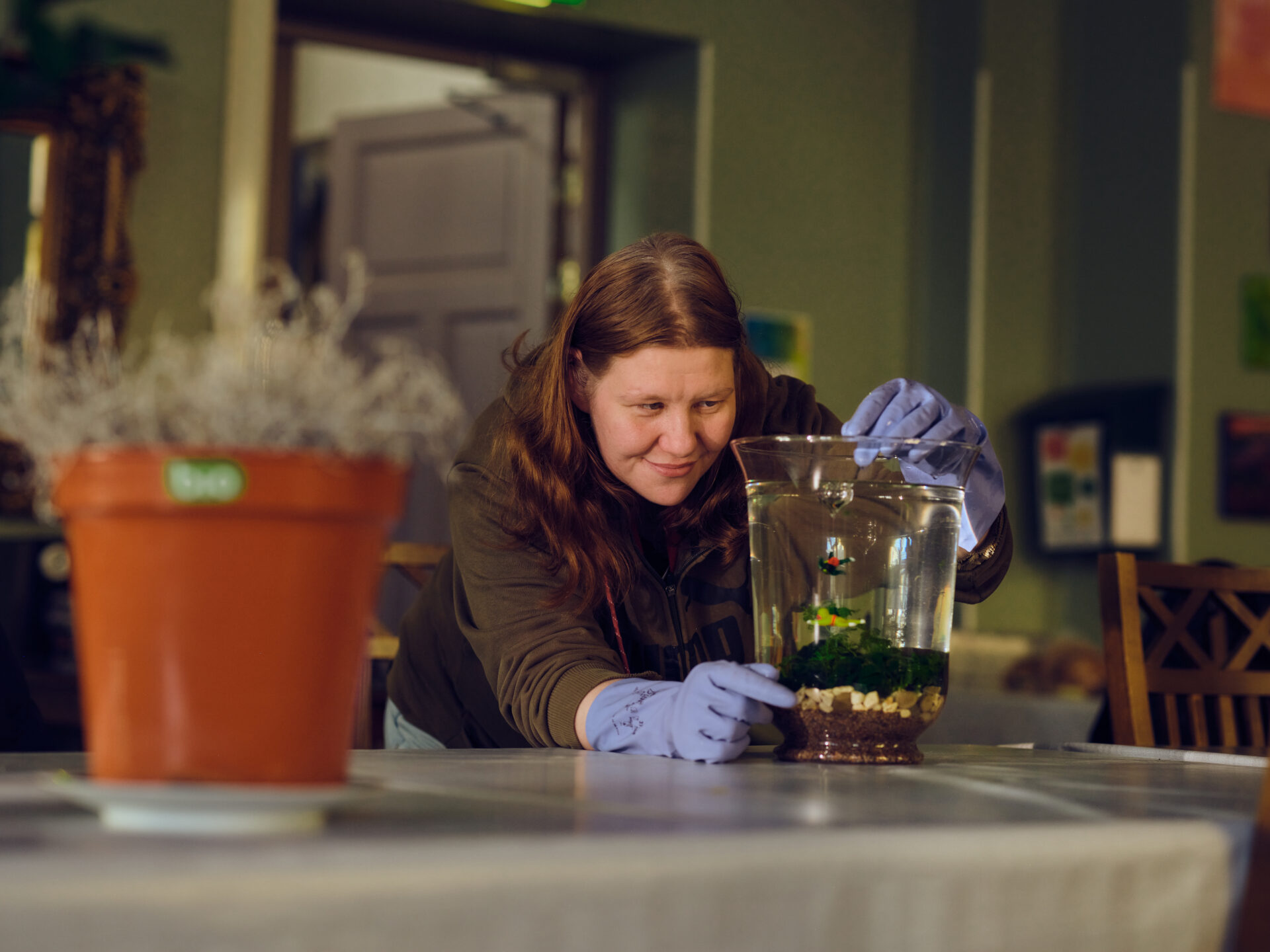 Pitkätukkainen hymyilevä nainen tutkii pientä lasiterraariota, jossa on kasveja.