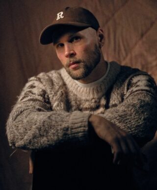 Lippalakkipäinen ja sänkipartainen mies istuu ilmeisesti hajareisin tuolilla niin, että hänen kätensä ovat tuolin selkämyksellä. Hänellä on paksu kuvioneulevillapaita.