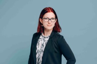 Polkkatukkainen, silmälasipäinen nainen, jolla on kuviollinen pusero ja bleiseri.